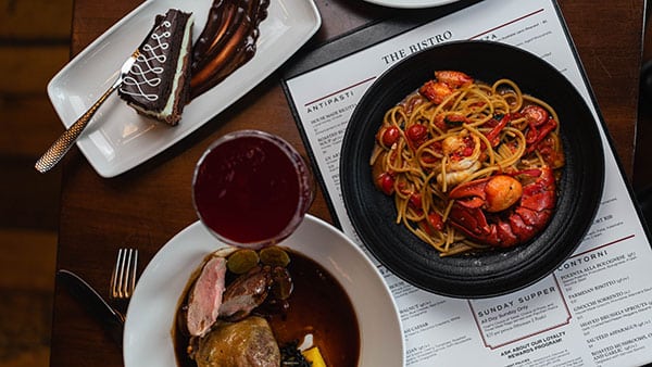 Flat-lay of gourmet dinner entrees including lobster pasta, Venetian duck, and chocolate dessert with a craft cocktail at The Bistro at Gervasi Vineyard.