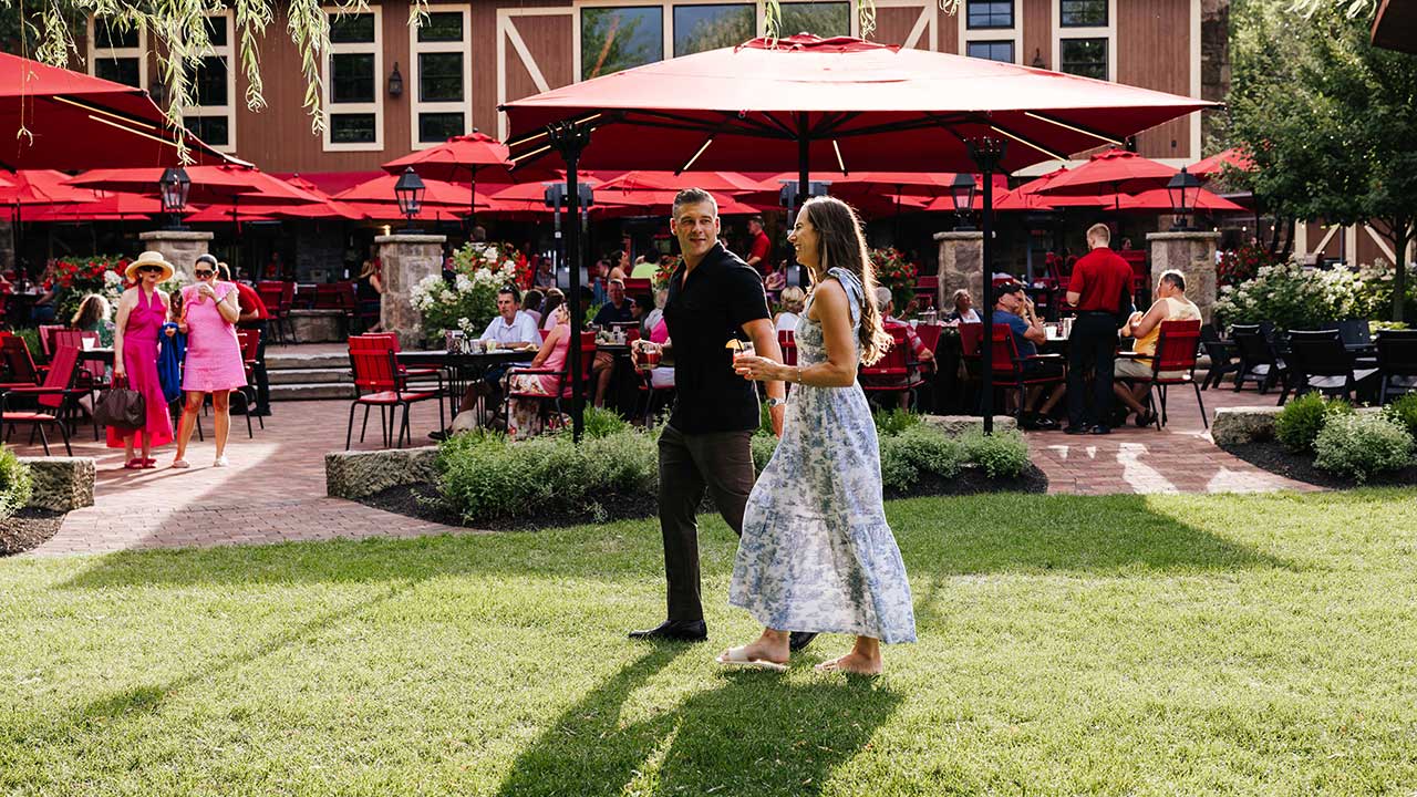 Guests dining al fresco under red umbrellas at The Piazza at Gervasi Vineyard, with views of the lake and Tuscan-style architecture.