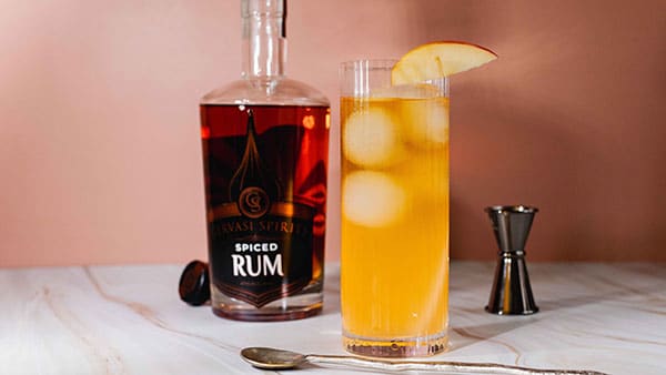 Gervasi Spirits Spiced Rum next to a tall apple rum cocktail with ice and a fresh apple garnish on a pink background.