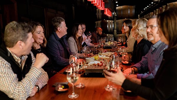 A large group enjoying a wine tasting event inside The Cave at Gervasi Vineyard, surrounded by red pendant lighting and wine barrels, creating a cozy and refined ambiance.