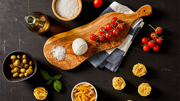 Rustic display of Italian cooking ingredients including fresh mozzarella, cherry tomatoes, pasta, olives, olive oil, and sea salt on a dark background