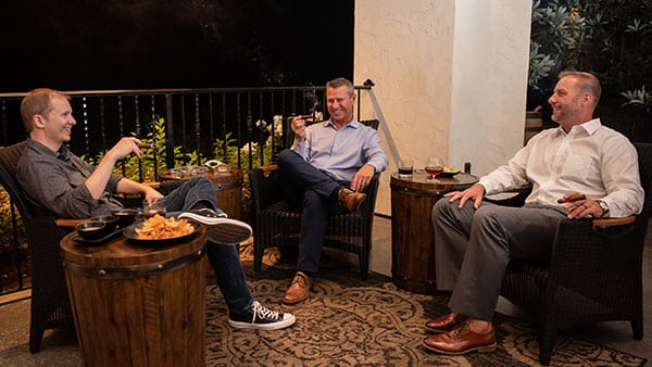 Three men enjoying cigars and bourbon in comfortable barrel-style chairs at Gervasi Vineyard’s outdoor cigar lounge.