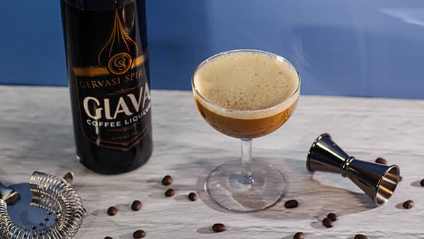Giava coffee liqueur from Gervasi Spirits beside a freshly shaken espresso martini with coffee beans and bar tools on a white surface