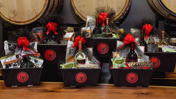 Assorted Gervasi Vineyard holiday gift baskets filled with wine bottles, gourmet treats, chocolates, and festive packaging, displayed in front of wine barrels at The Marketplace boutique