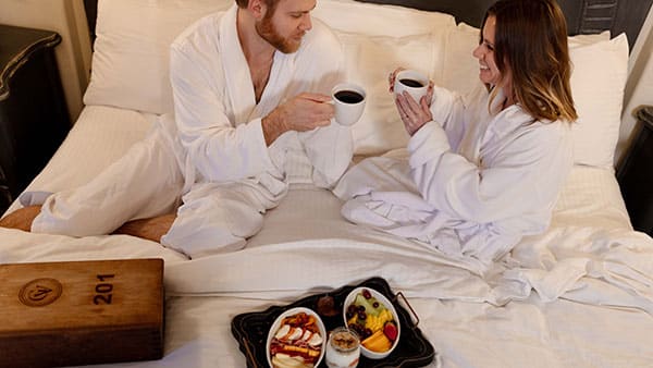 Couple enjoying breakfast in bed wearing white robes at Gervasi Vineyard’s luxury villa suite.