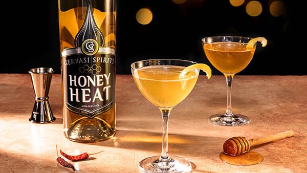 Gervasi Spirits Honey Heat bottle with spicy honey cocktails garnished with lemon, chili peppers, and honey dipper on bar surface