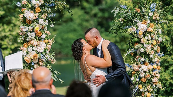 Bride and groom sharing a kiss during an outdoor wedding ceremony framed by floral pillars at Gervasi Vineyard.