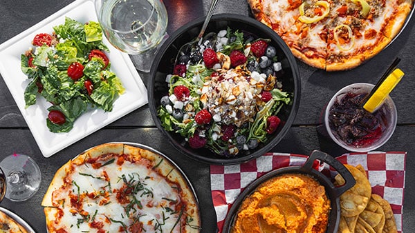 Overhead view of artisan pizzas, fresh salad, hummus appetizer, and drinks served at Gervasi Vineyard outdoor dining
