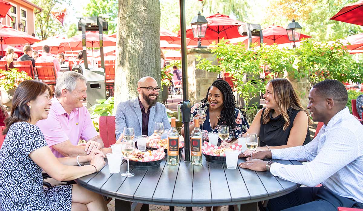 Friends sharing wine and Italian cuisine on the Piazza patio at Gervasi Vineyard under red umbrellas.