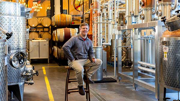 Gervasi Spirits distiller in the distillery, seated among copper stills, barrels, and distillation equipment.
