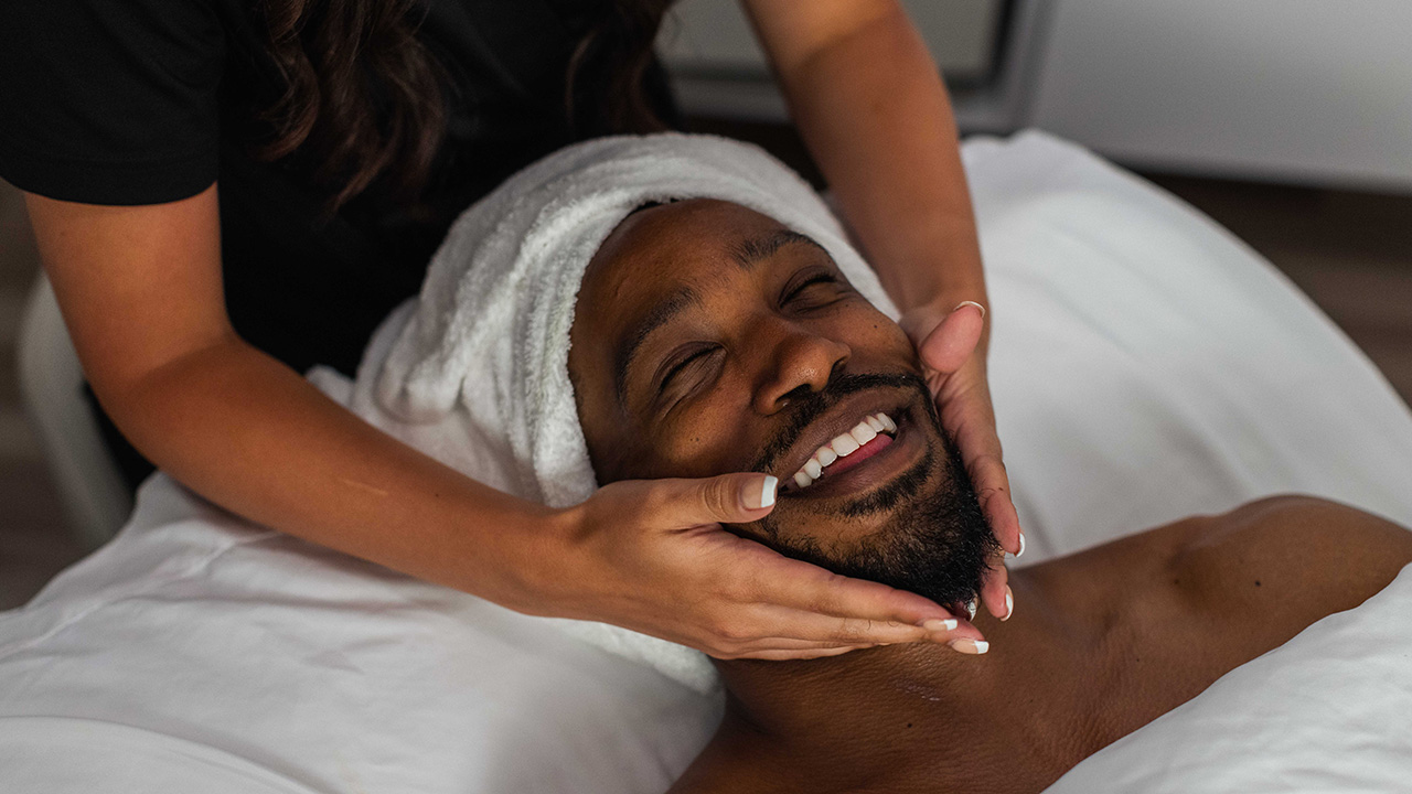 A man at The Spa at Gervasi Vineyard in Canton, Ohio enjoys a relaxing facial massage, wrapped in a white towel with a serene, smiling expression.