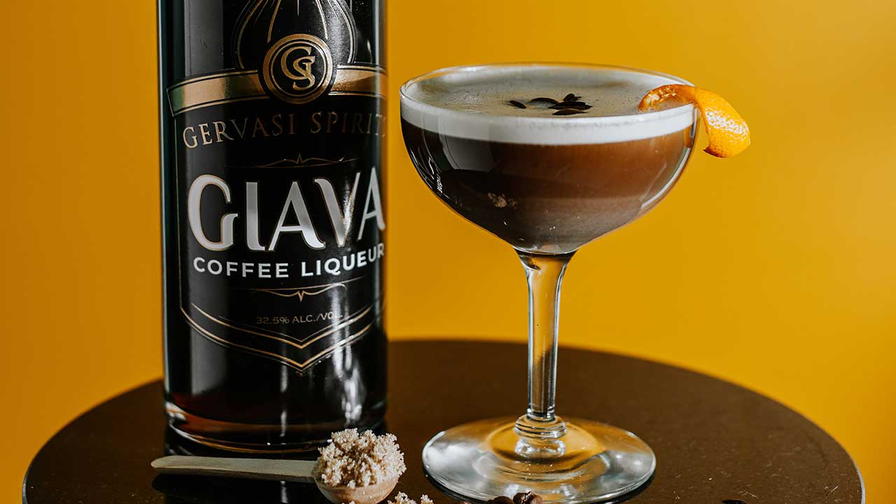 Gervasi Spirits Giava coffee liqueur bottle next to a rich espresso martini garnished with coffee beans and orange peel on a bold yellow background.