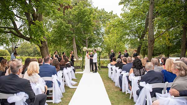Outdoor wedding ceremony at Gervasi Vineyard under a canopy of trees, with the bride and groom exchanging vows in front of guests seated along a white aisle runner