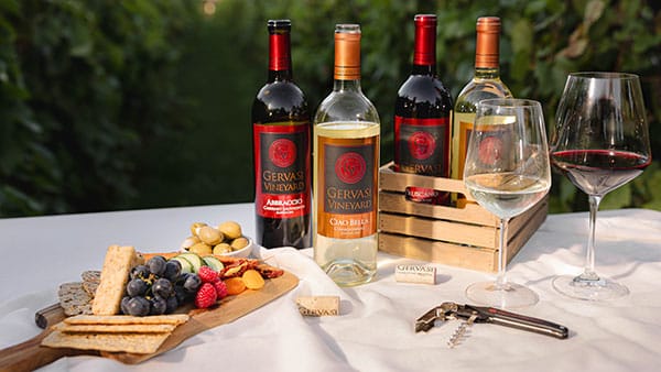Gervasi Vineyard wine bottles with red and white wine glasses, corkscrew, and gourmet charcuterie board in vineyard setting