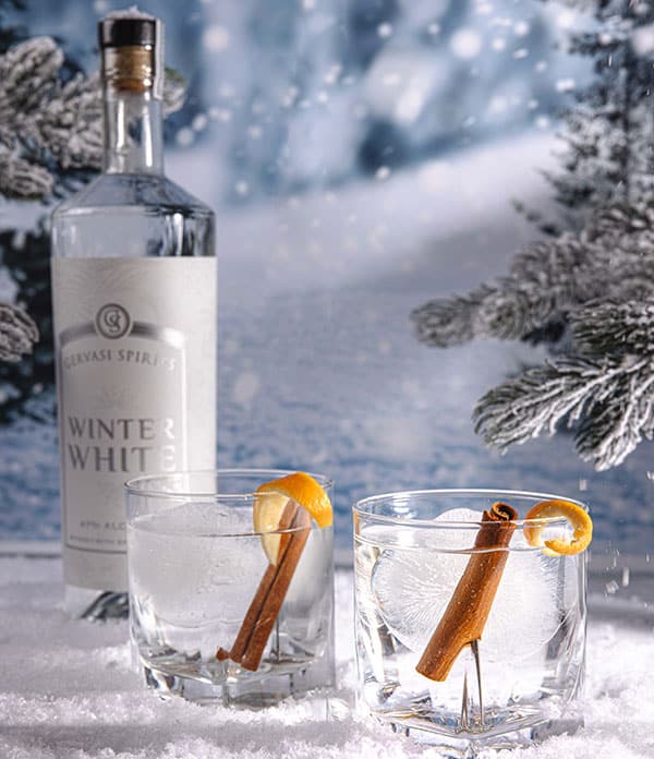 Winter White cocktails garnished with orange peel and cinnamon sticks, set in a snowy landscape with Gervasi Spirits bottle in background